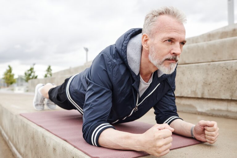 Sporty determined elderly man holding plank position on yoga mat strengthening core muscles during everyday workout at city park